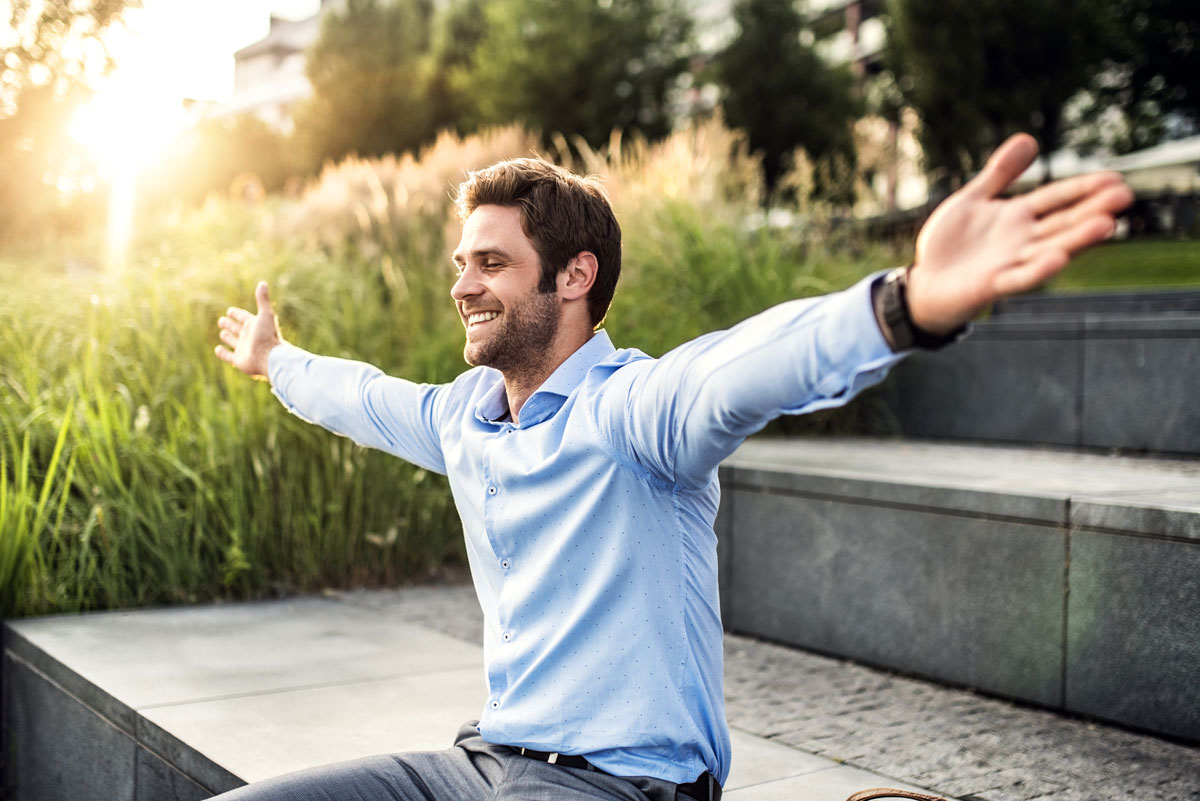 A happy young businessman with arms stretched outdoors at sunset.